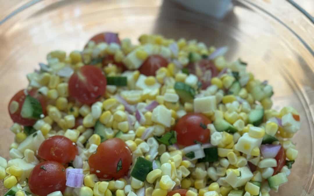Colorful Mediterranean corn and tomato salad with cucumbers, red onion, basil, and crumbled Asiago cheese in a white serving bowl