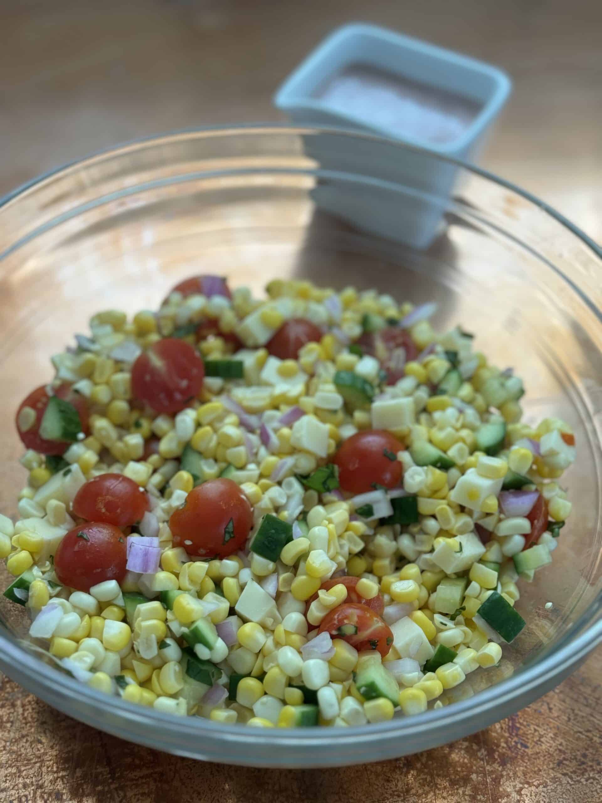 Colorful Mediterranean corn and tomato salad with cucumbers, red onion, basil, and crumbled Asiago cheese in a white serving bowl